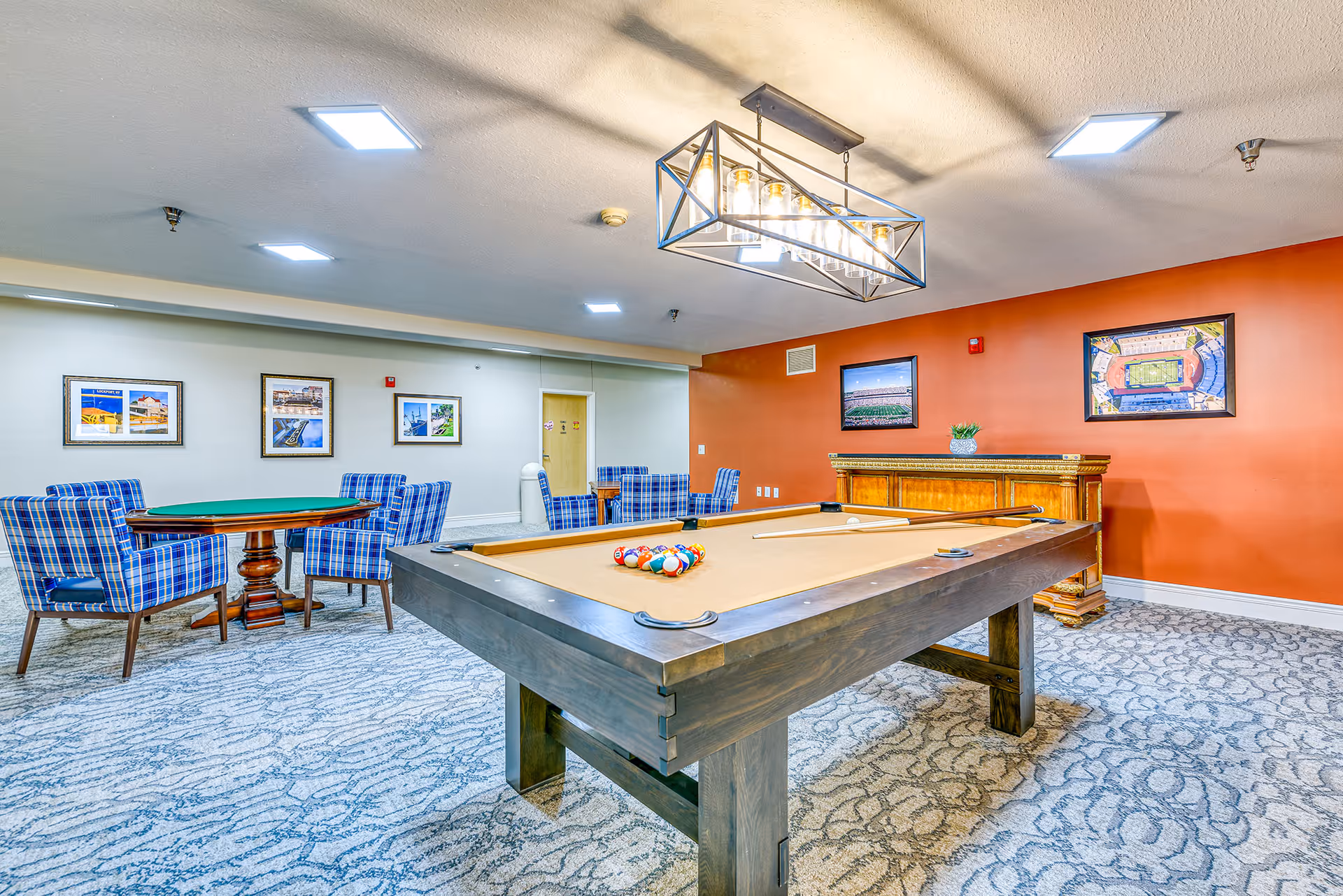 A bright game room with a pool table in the foreground, plaid-upholstered chairs around card tables, and framed art on warm-colored walls.