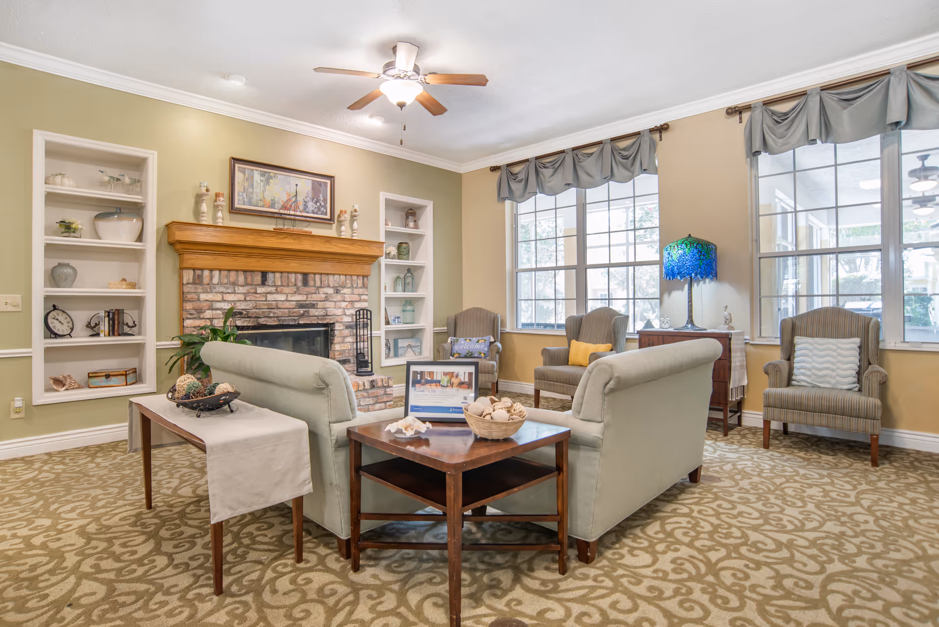 Communal living room with sofas and armchairs arranged around a brick fireplace beneath built-in shelves and large windows.