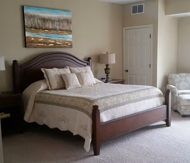 A neatly made bed with a wooden headboard and footboard in a bedroom. The bed has a quilted bedspread and several decorative pillows. There is a nightstand with a lamp and an alarm clock next to the bed. A cushioned armchair with a floral pattern is positioned near a closed door. A landscape painting hangs on the wall above the bed.