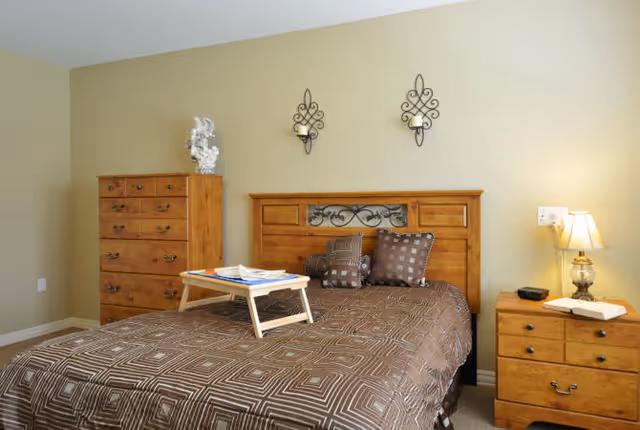 A bedroom with a wooden bed frame and matching wooden dresser and nightstand. The bed is made with a brown patterned bedspread and several decorative pillows. There is a small wooden tray on the bed with some papers and a book. Two decorative wall sconces are mounted above the headboard. A lamp and an open book are on the nightstand.