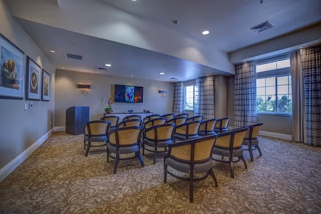 A small conference or presentation room with rows of chairs facing a wall-mounted flat screen TV. The room has patterned carpet, beige walls with framed artwork, two windows with curtains allowing natural light, and a podium at the front left corner.