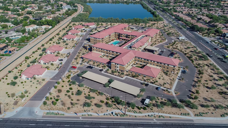 Aerial view of a senior living complex with interconnected red‑roofed buildings, covered parking, landscaped grounds, and a nearby lake.