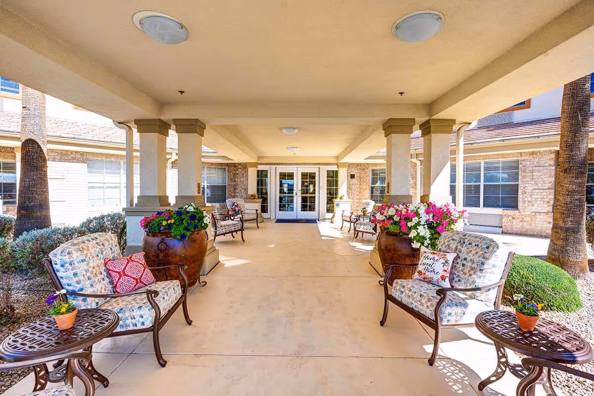 Covered outdoor seating area at Sun City West Assisted Living with cushioned chairs, small tables, large flower pots with colorful flowers, and a view of the building entrance with glass doors.