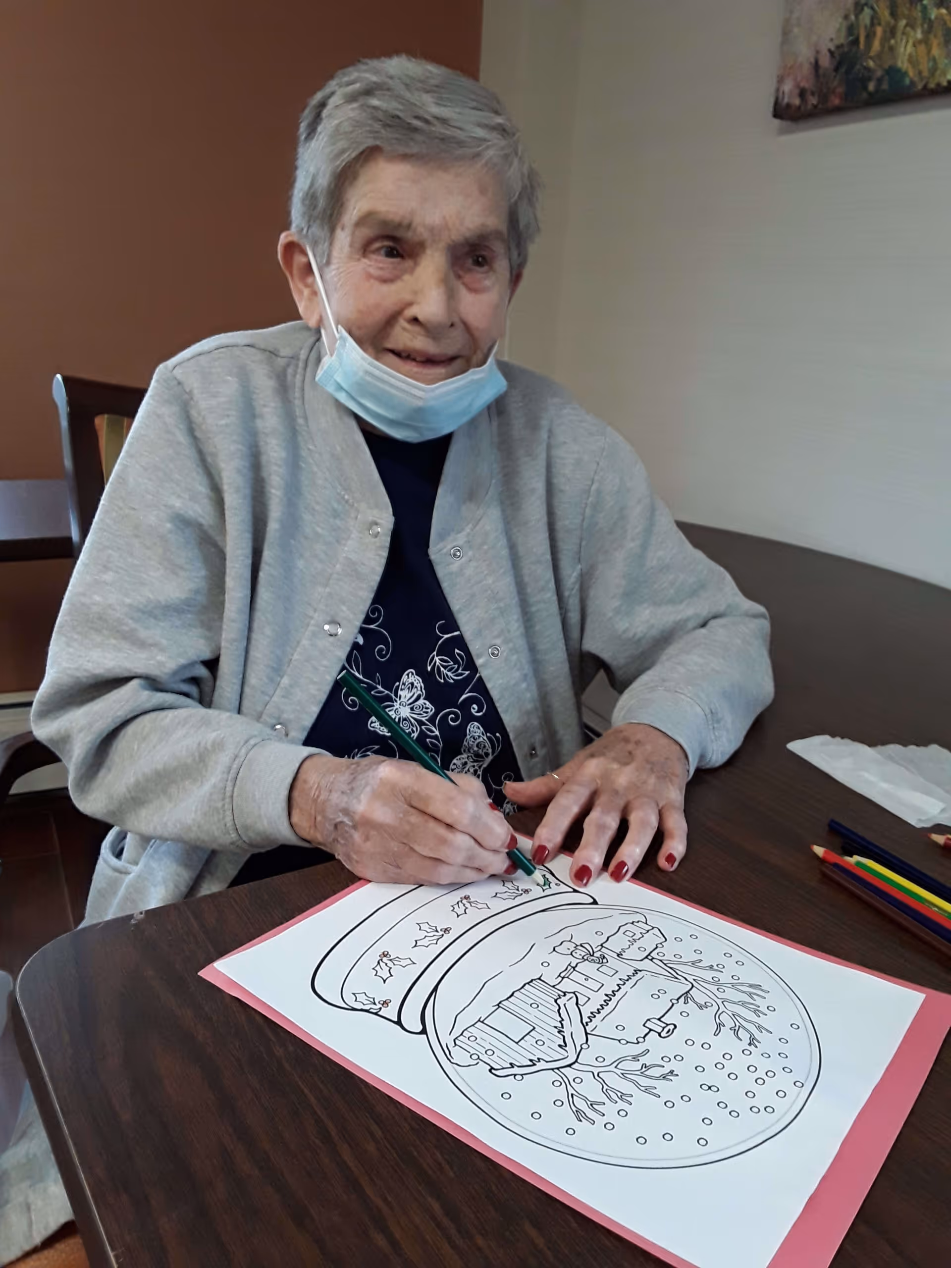 An elderly woman wearing a gray cardigan and a partially lowered face mask is sitting at a wooden table, coloring a winter-themed drawing with colored pencils. The drawing features a snow globe with a house and trees inside. The woman is smiling gently and appears engaged in the activity.