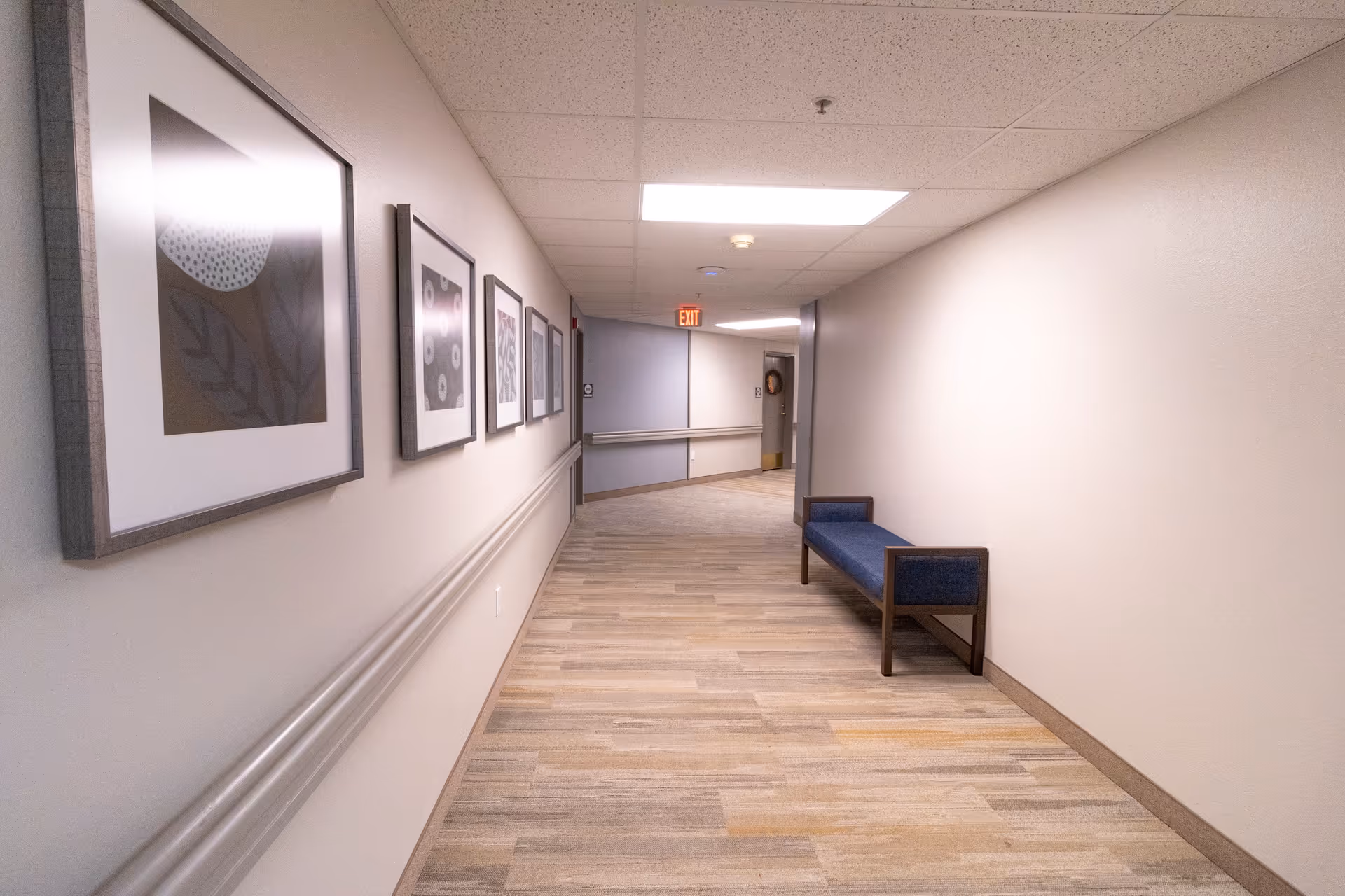 A well-lit indoor hallway with light-colored walls and wood-patterned flooring. On the left wall, there are several framed abstract art pieces. A blue cushioned bench with wooden arms is placed against the right wall. The hallway leads to a door with an exit sign above it.