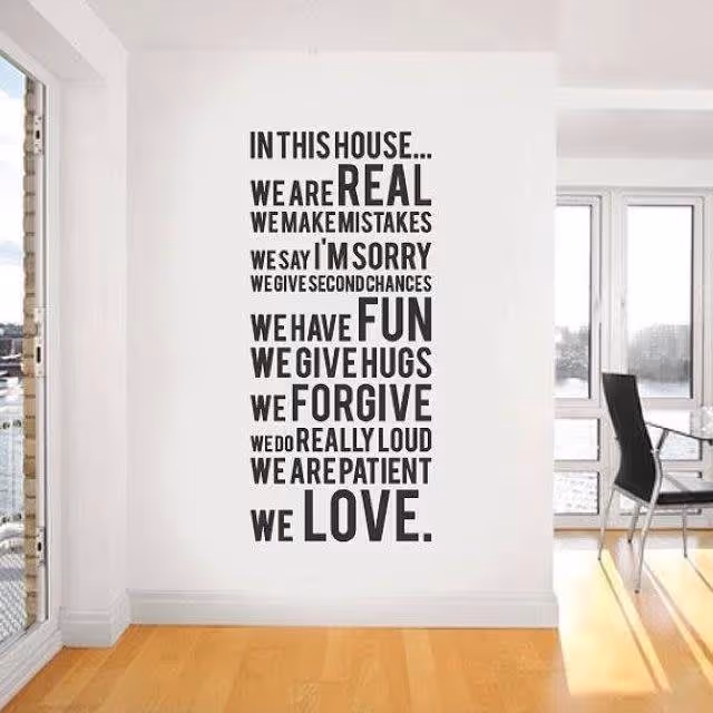 A white wall with a large black text decal that reads: 'IN THIS HOUSE... WE ARE REAL WE MAKE MISTAKES WE SAY I'M SORRY WE GIVE SECOND CHANCES WE HAVE FUN WE GIVE HUGS WE FORGIVE WE DO REALLY LOUD WE ARE PATIENT WE LOVE.' The room has light wooden flooring and large windows letting in natural light. A black chair and a glass table are partially visible on the right side.
