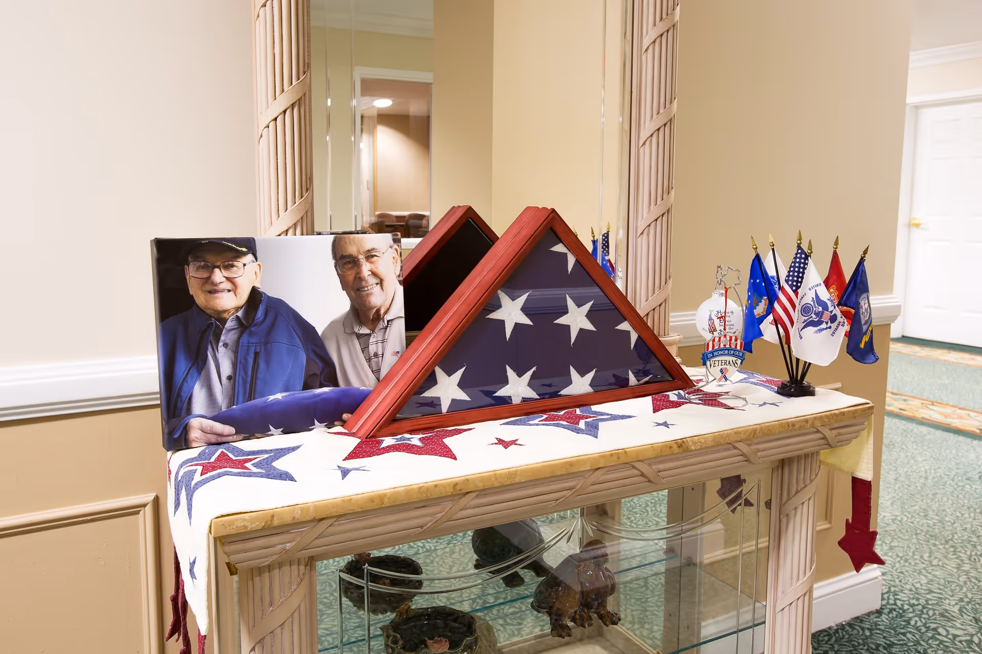 A memorial display on a wooden table with a patriotic star-patterned cloth, featuring a folded American flag in a triangular wooden case, a photo of two elderly men, and several small flags representing different branches of the U.S. military. The setting appears to be an indoor hallway with beige walls and a green carpet.