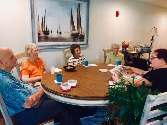 Five residents seated around a round wooden table in a communal room with cups and snacks while a staff member reads aloud.