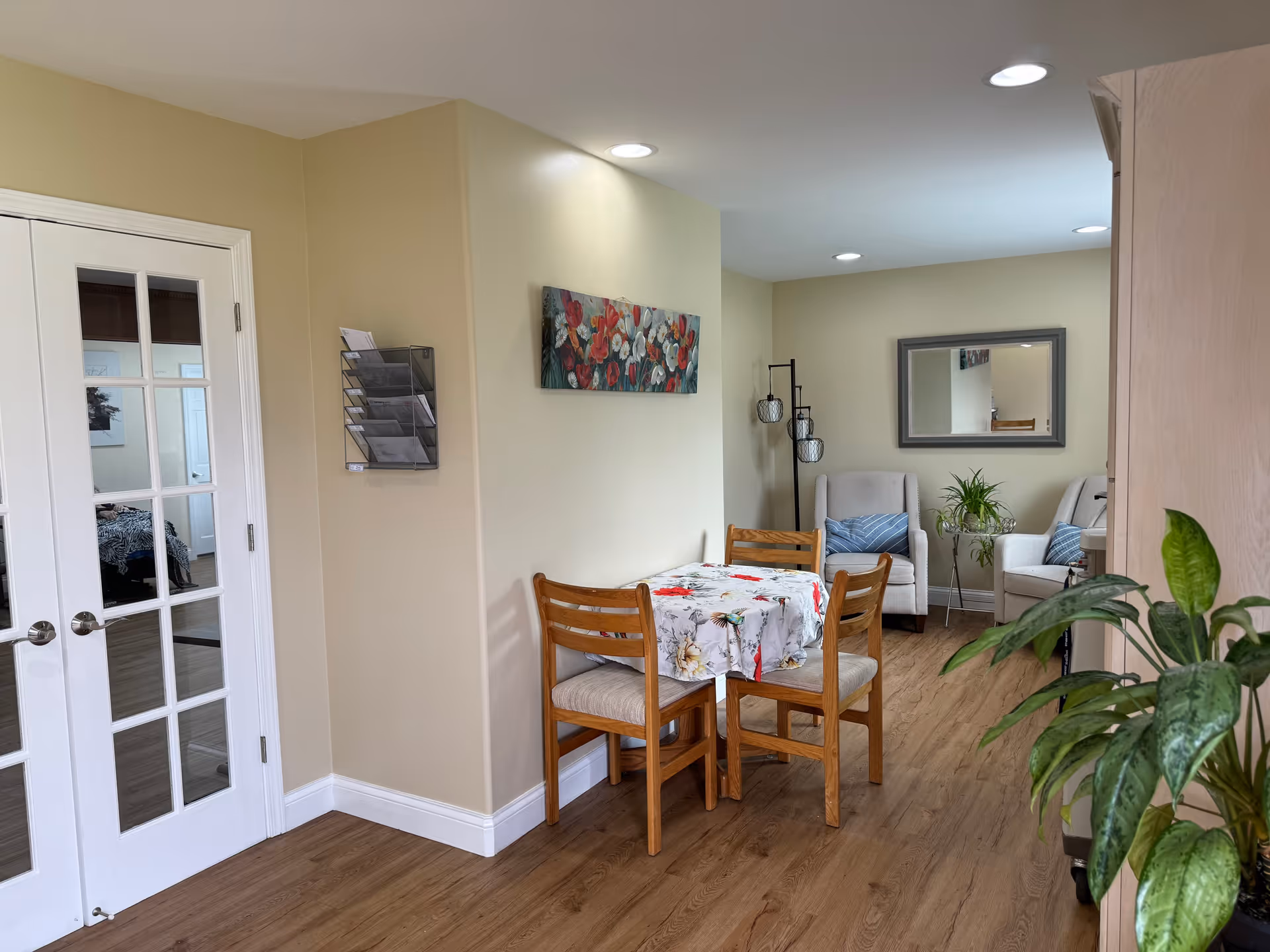 A cozy interior space featuring a small wooden dining table with a floral tablecloth and three wooden chairs. In the background, there are two cushioned armchairs with blue pillows, a floor lamp with three lights, a small side table with a plant, and a large wall mirror. The floor is wood, and the walls are painted light beige. To the left, there is a set of white French doors with glass panes, and a wall-mounted brochure holder is visible.
