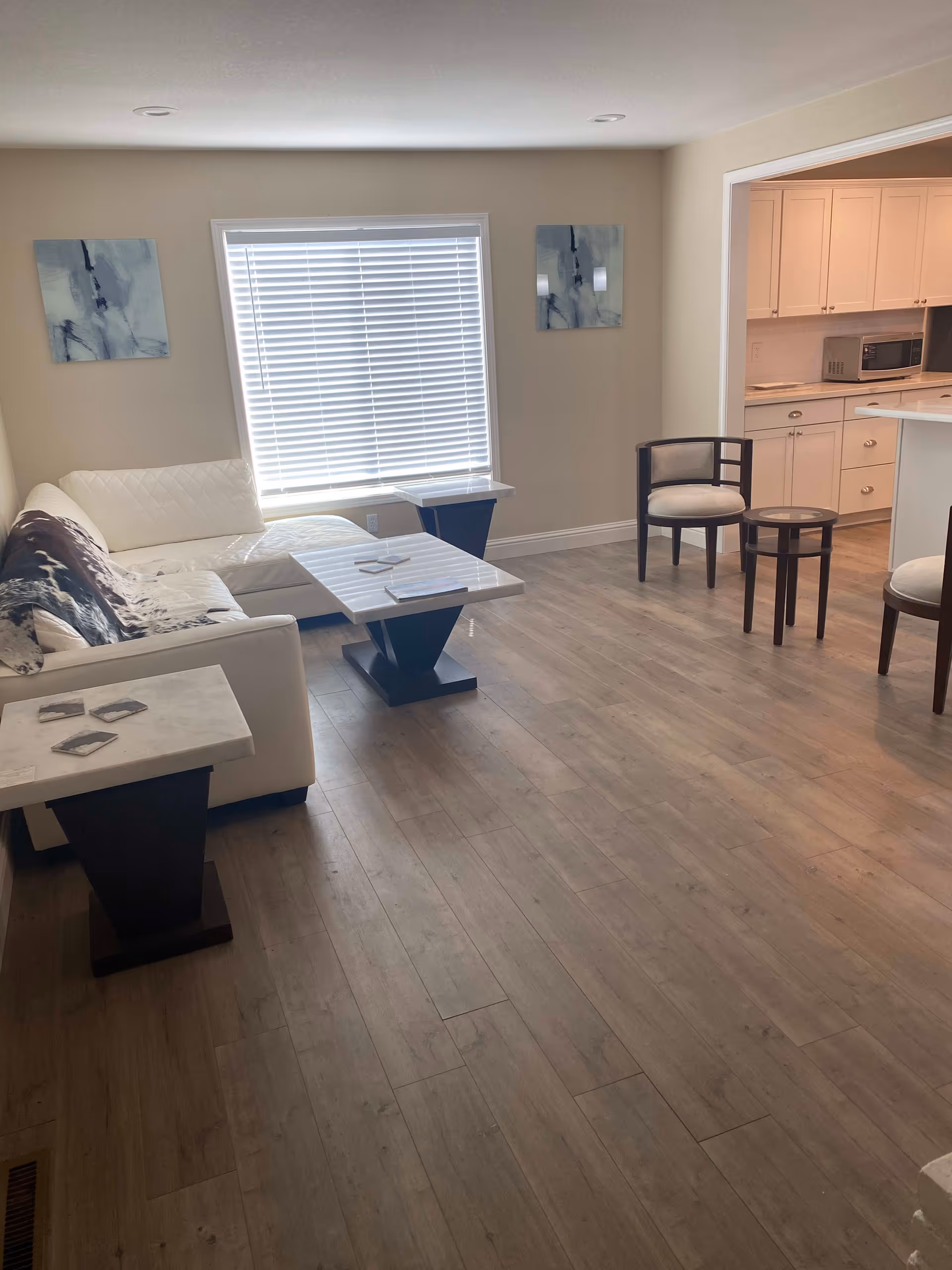 A bright living room with light wood flooring, a white sectional sofa with a black and white throw, two marble-top tables, two wooden chairs with light cushions, and a large window with white blinds. The room opens into a kitchen area with white cabinets and a microwave.