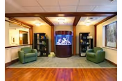 A cozy interior common area featuring two green armchairs positioned on either side of a large cylindrical aquarium filled with colorful coral and fish. The space has wooden flooring, a green area rug, bookshelves with books and decorative items, and soft lighting from ceiling fixtures. A large window on the right side lets in natural light.