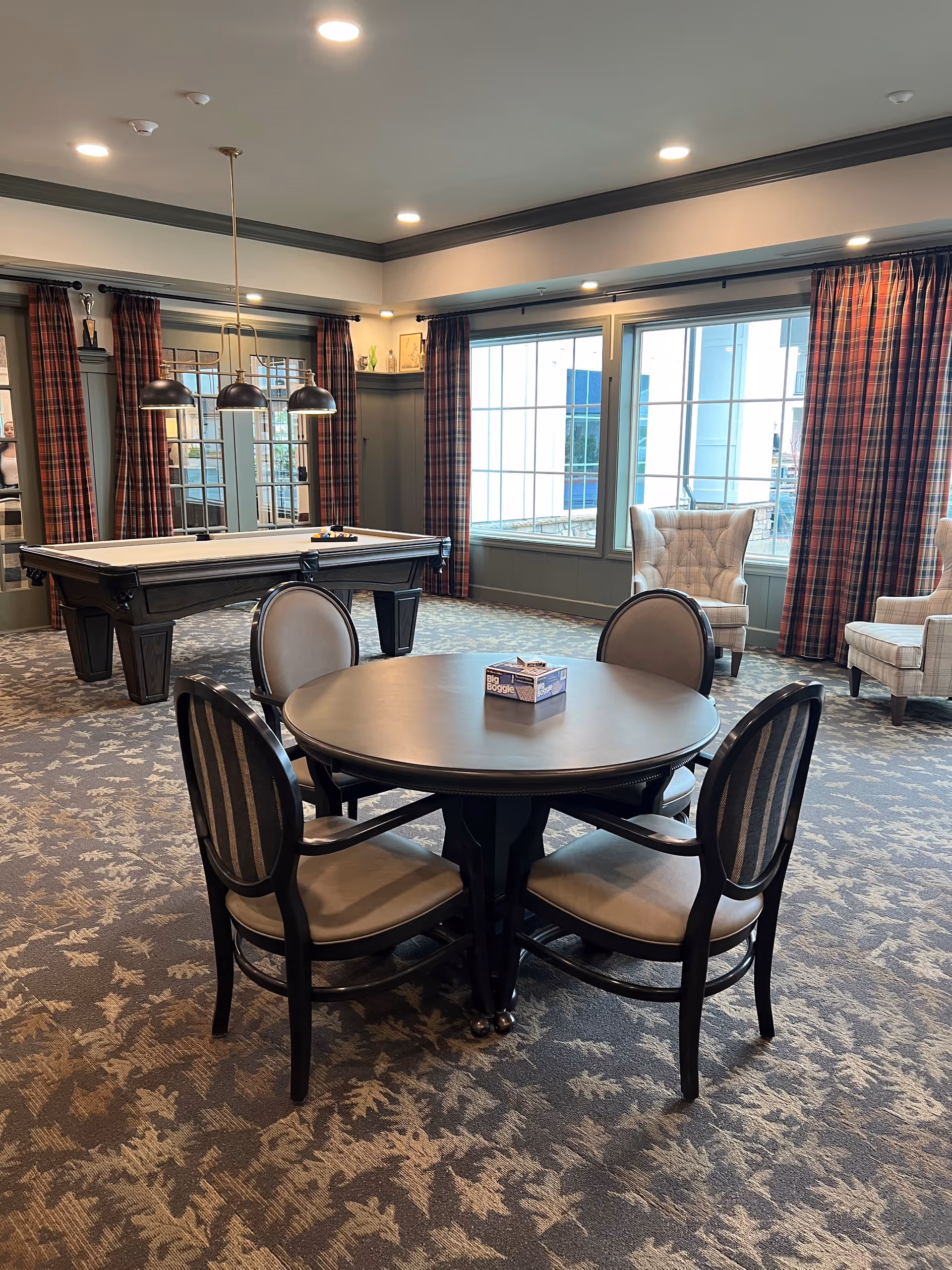 A cozy communal lounge with a round table and four chairs, a pool table, armchairs, and large windows with plaid curtains.