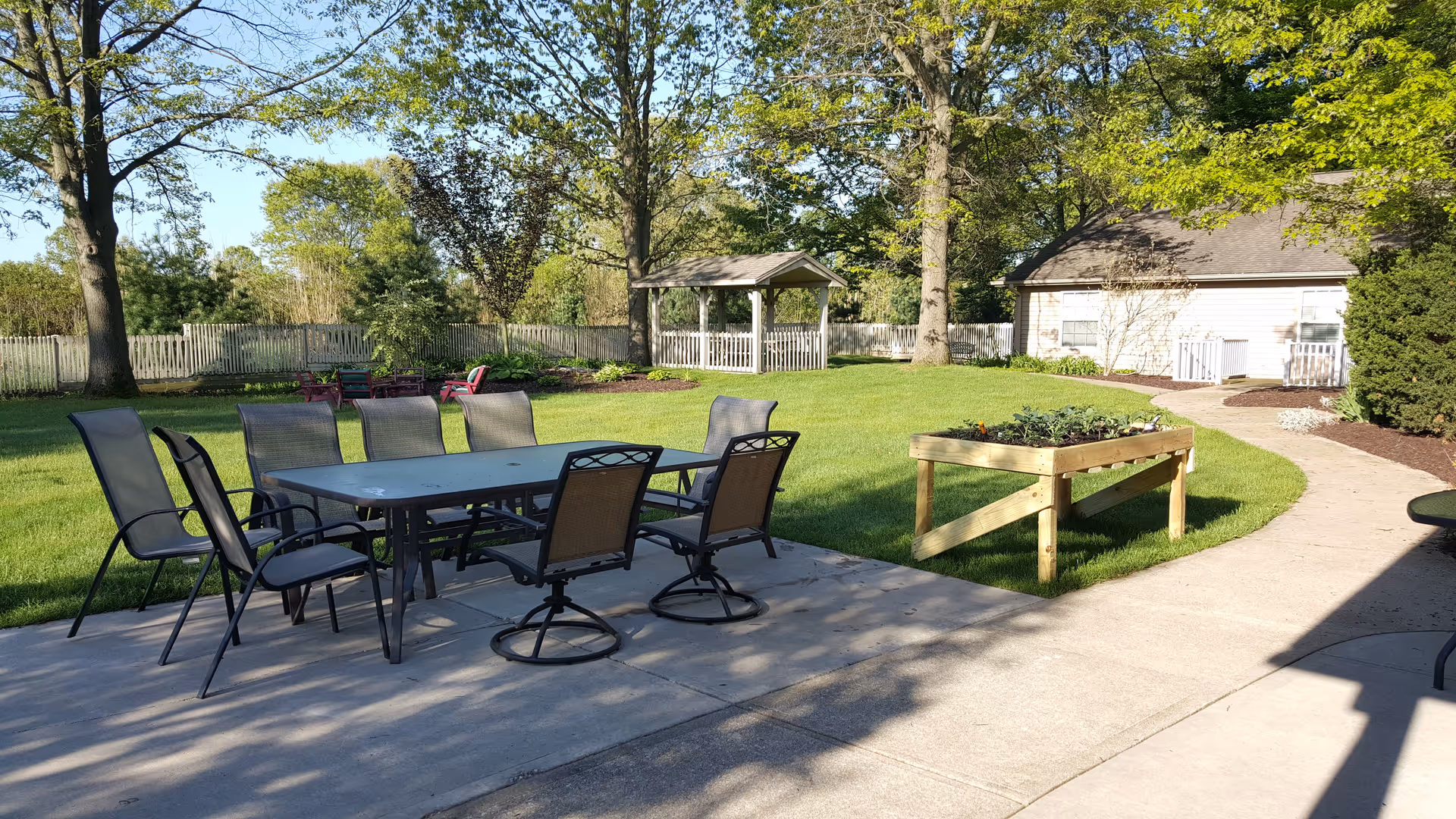 Outdoor patio area with a rectangular table surrounded by eight chairs on a concrete surface. In the background, there is a grassy lawn with trees, a wooden gazebo, a raised garden bed, and a light-colored building with a sloped roof. The scene is bright and sunny with clear skies.