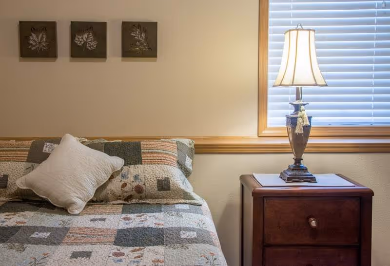 A cozy bedroom corner featuring a bed with a patchwork quilt and a white pillow, a wooden nightstand with a decorative lamp, and a window with white blinds. Three small framed leaf artworks hang on the beige wall above the bed.