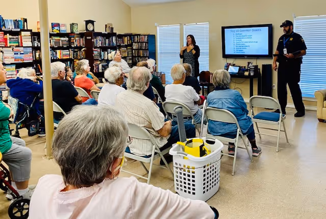 A group of elderly people seated on folding chairs in a room with bookshelves, attentively watching a presentation given by a man and a woman. The woman is using sign language, and a screen displays a slide titled 'Top 10 Common Scams'.