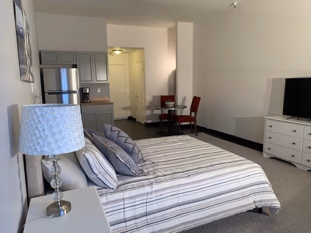 A spacious bedroom with a bed covered in striped bedding and multiple pillows. Next to the bed is a white nightstand with a decorative lamp. In the background, there is a small kitchenette with a refrigerator, cabinets, and a coffee maker. A small round dining table with two red chairs is placed near the kitchenette. A white dresser with a flat-screen TV on top is positioned against the opposite wall. The room has neutral-colored walls and carpeted flooring.