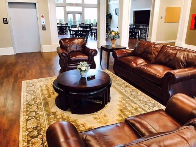 A cozy living room area with brown leather sofas and armchairs arranged around a round wooden coffee table with a small flower arrangement. The room has wooden flooring and a patterned area rug. In the background, there is an elevator on the left and a dining area with tables and chairs visible through an open doorway.