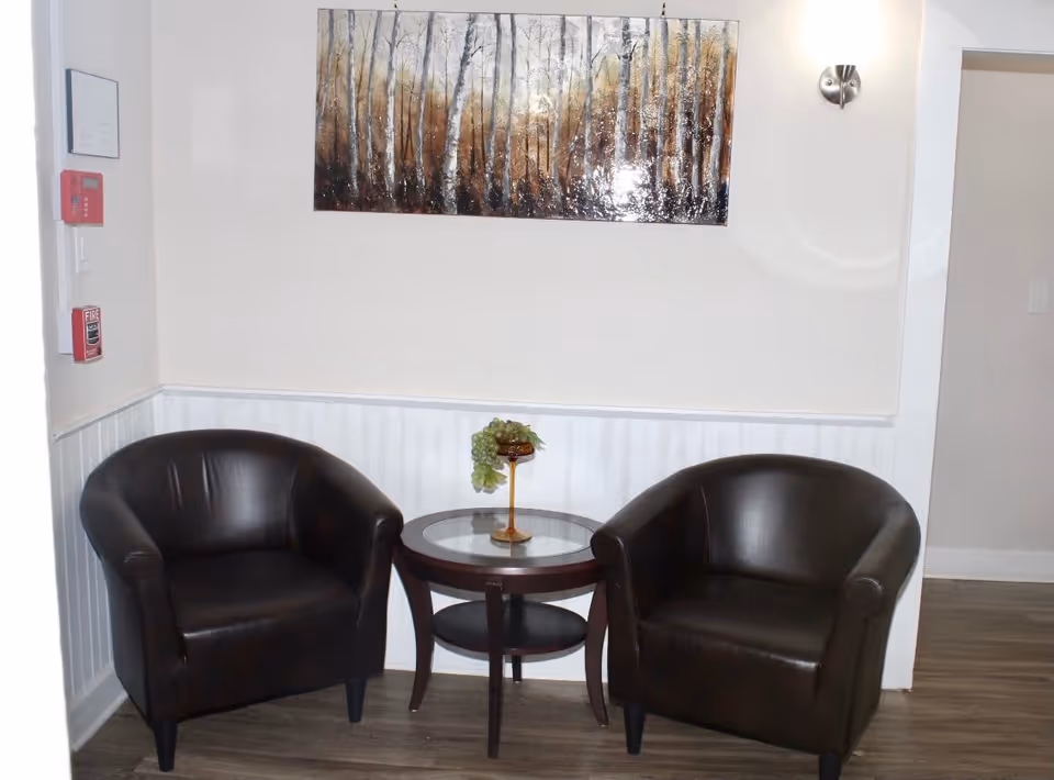 A small seating area with two black leather armchairs facing each other and a round glass-top wooden table between them. On the table is a decorative item with green grapes. Above the seating area is a wall-mounted painting depicting a forest of birch trees. The walls are light-colored with white wainscoting, and there is a wall sconce light fixture on the right side.