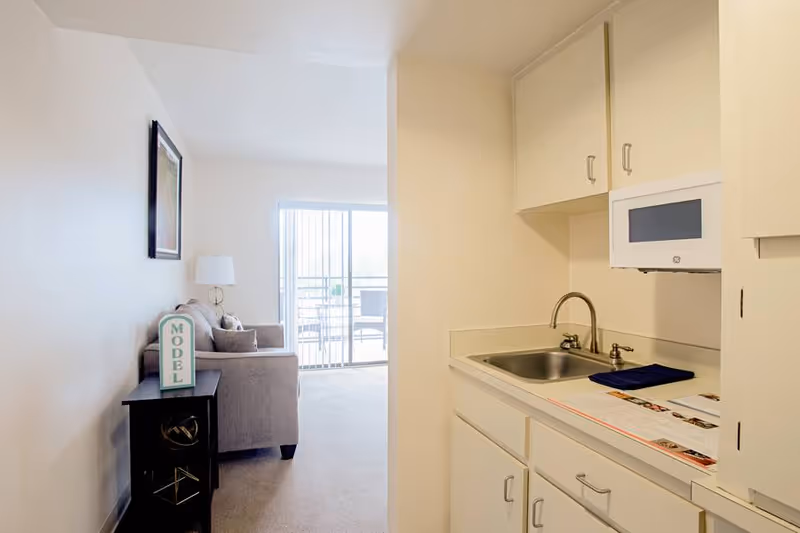 Small apartment interior showing a kitchenette with sink and cabinets opening to a living area with a sofa and sliding glass door to a balcony.