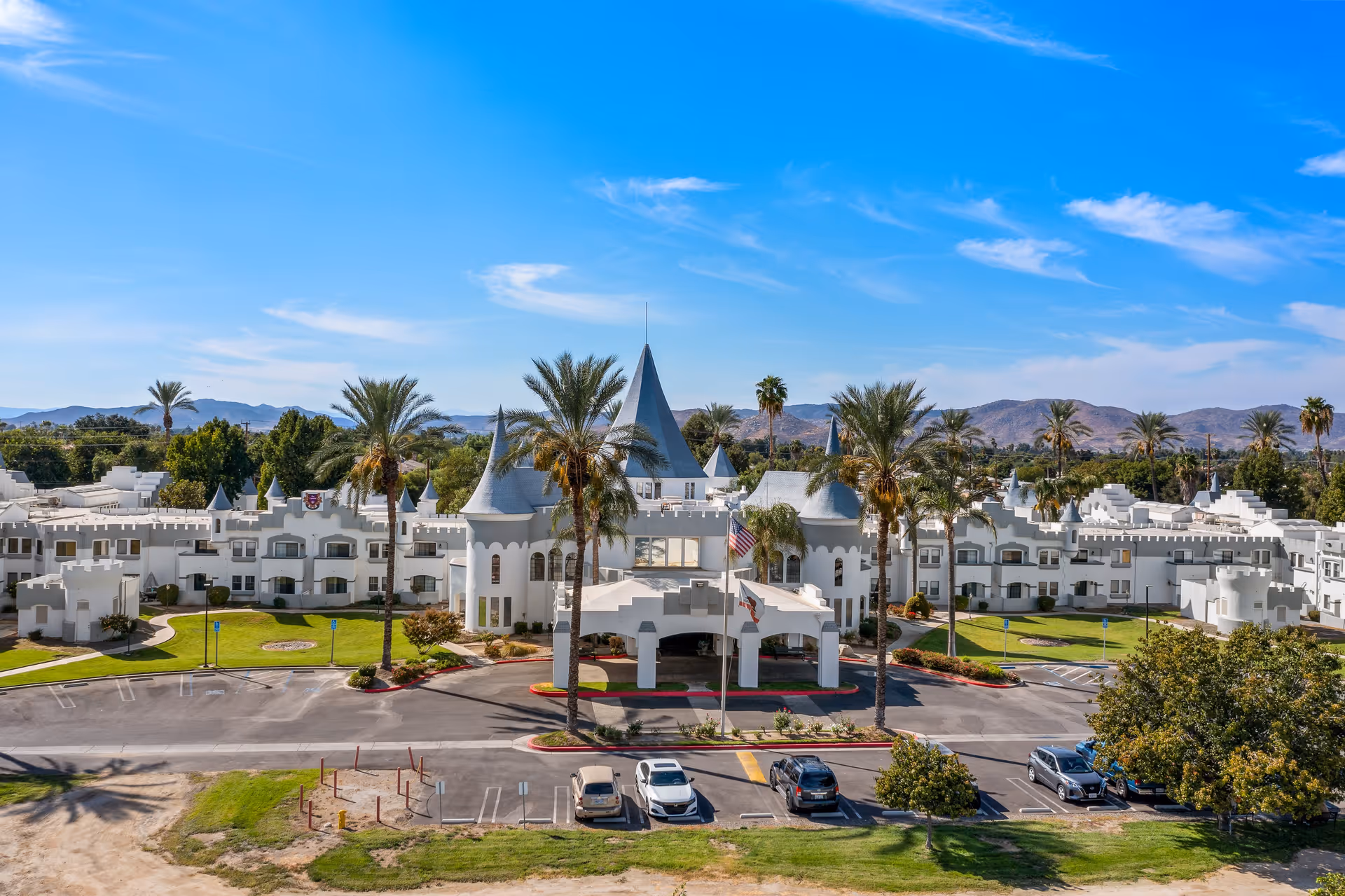 Front exterior view of Camelot - A Provincial Senior Living Community, a large white building with castle-like architecture including turrets and battlements, surrounded by palm trees and greenery under a blue sky with some clouds. There is a parking lot with several cars in front of the building.