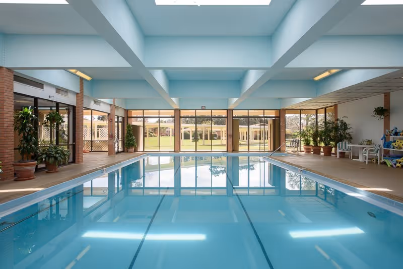 Indoor swimming pool area with clear blue water, surrounded by large windows letting in natural light. There are several potted plants around the pool and a seating area with chairs and pool toys on the right side. The ceiling has beams and skylights.