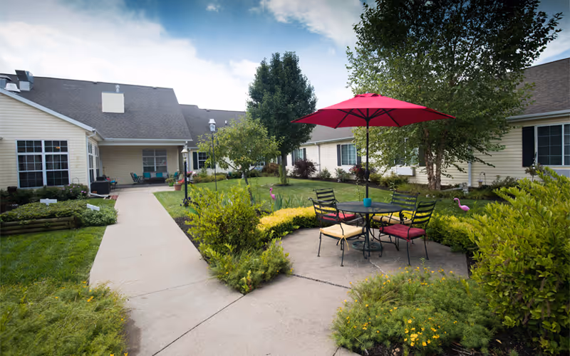 Outdoor courtyard area of a senior living facility with a concrete walkway, green bushes, trees, and a round glass table with four chairs under a red umbrella. The buildings surrounding the courtyard have beige siding and multiple windows.