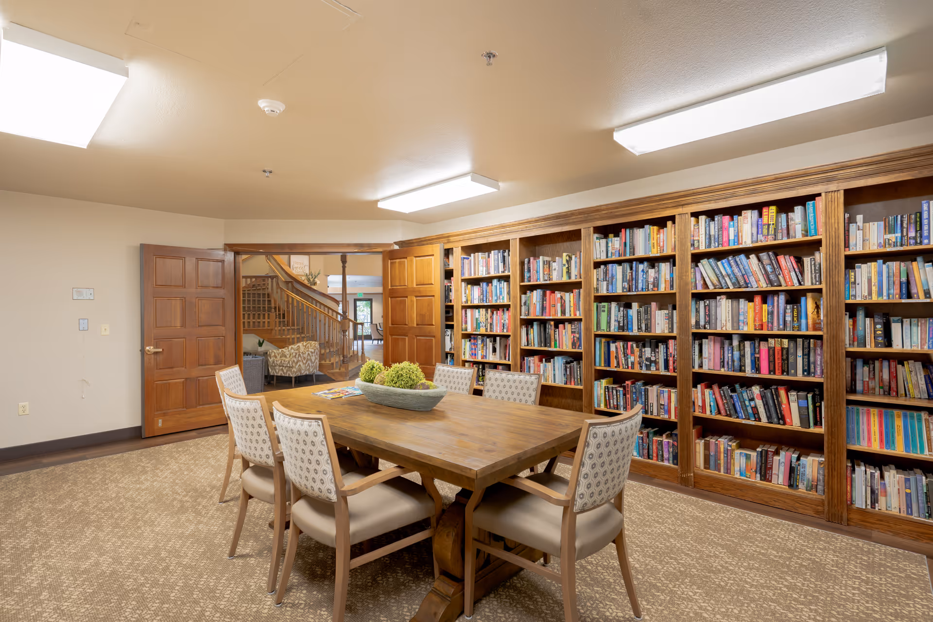 A cozy library room with wooden bookshelves filled with books along one wall. In the center, there is a wooden table with six cushioned chairs around it. A decorative bowl with green plants is placed on the table. The room has beige walls and carpeted floor. Double wooden doors are open, showing a glimpse of a staircase and seating area in the adjacent space.