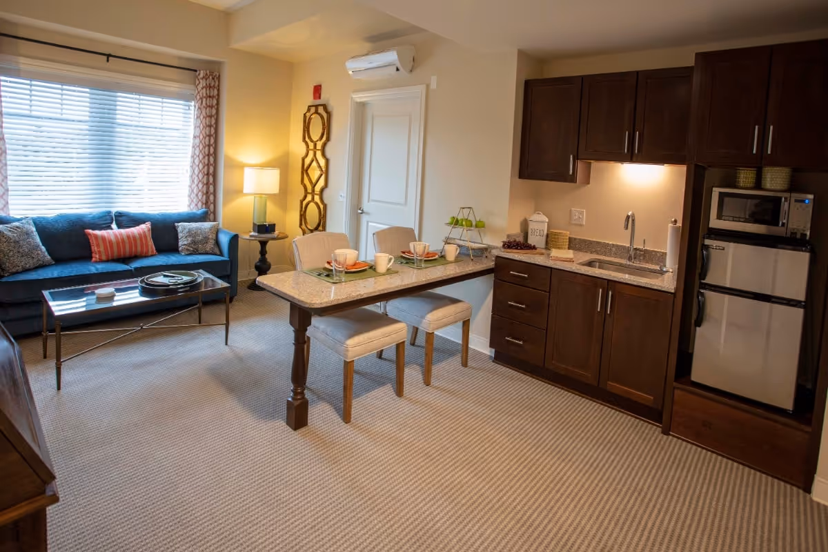 Open living area with a sofa and coffee table, a dining-style island with stools, and a kitchenette with dark cabinets and a mini-fridge.