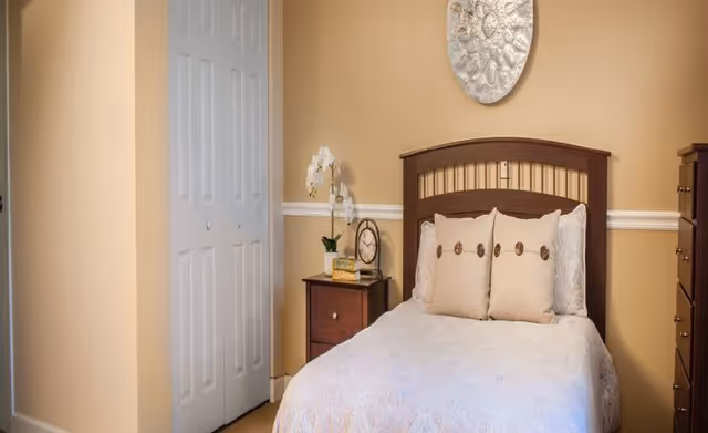 Cozy single bedroom with a wooden headboard, nightstand holding a clock and orchid, closet doors, and beige walls.