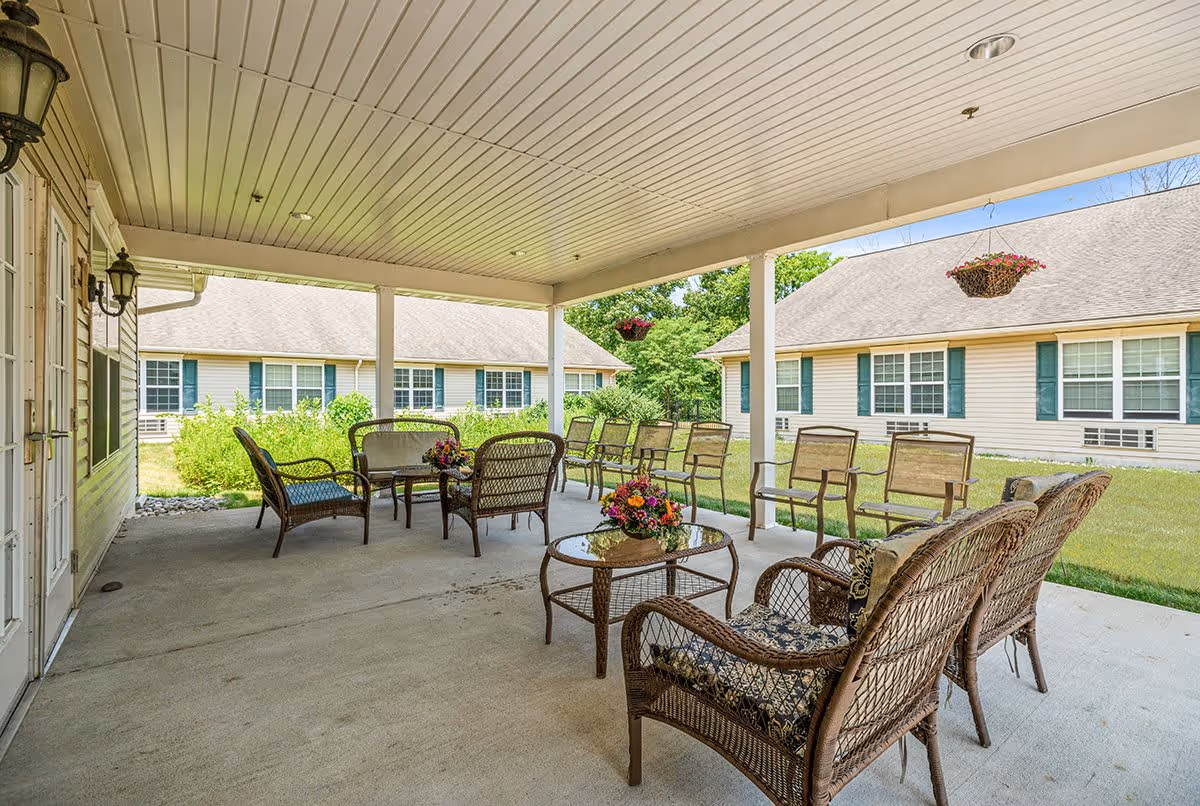 Covered outdoor patio area with wicker chairs and tables arranged for seating. The patio overlooks a grassy courtyard with two single-story buildings featuring beige siding and green shutters. Hanging flower baskets are suspended from the patio ceiling.