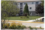 Front exterior of a multi-story apartment building with lawn, trees, and a sidewalk.