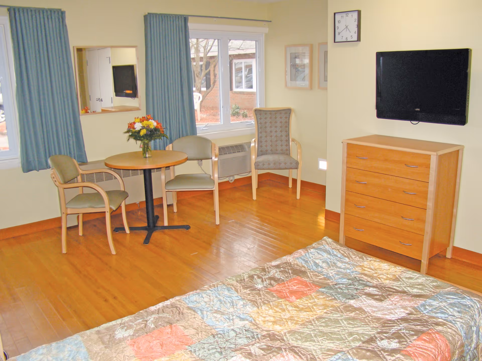 A bright room with wooden flooring featuring a bed with a colorful quilt in the foreground, a wooden dresser with a flat-screen TV mounted above it on the wall, a round table with a vase of flowers, and three chairs near two windows with blue curtains. A wall clock and framed pictures are also visible.