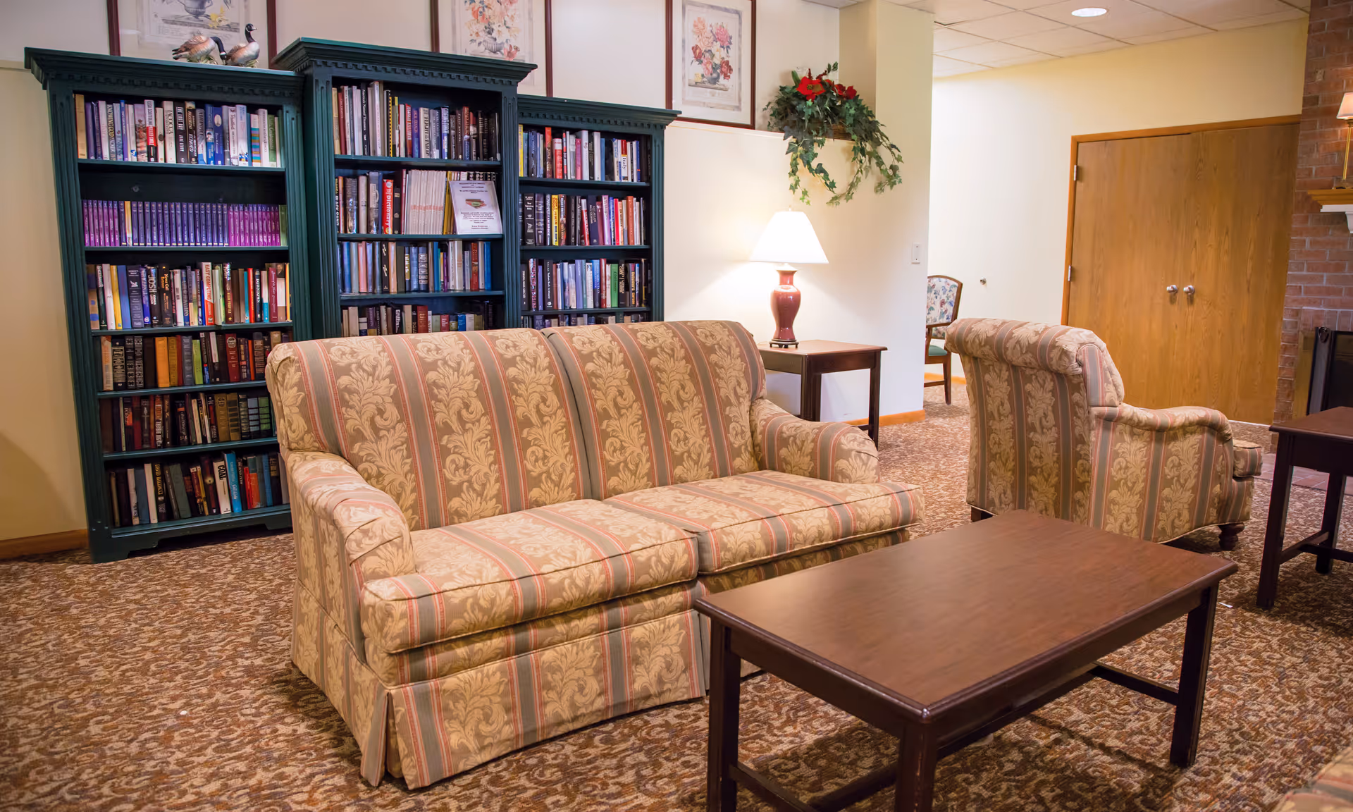 A cozy living room area with a patterned sofa and armchair, a wooden coffee table, a side table with a lamp, a green bookshelf filled with books, framed floral artwork on the wall, and a decorative plant hanging on the wall.