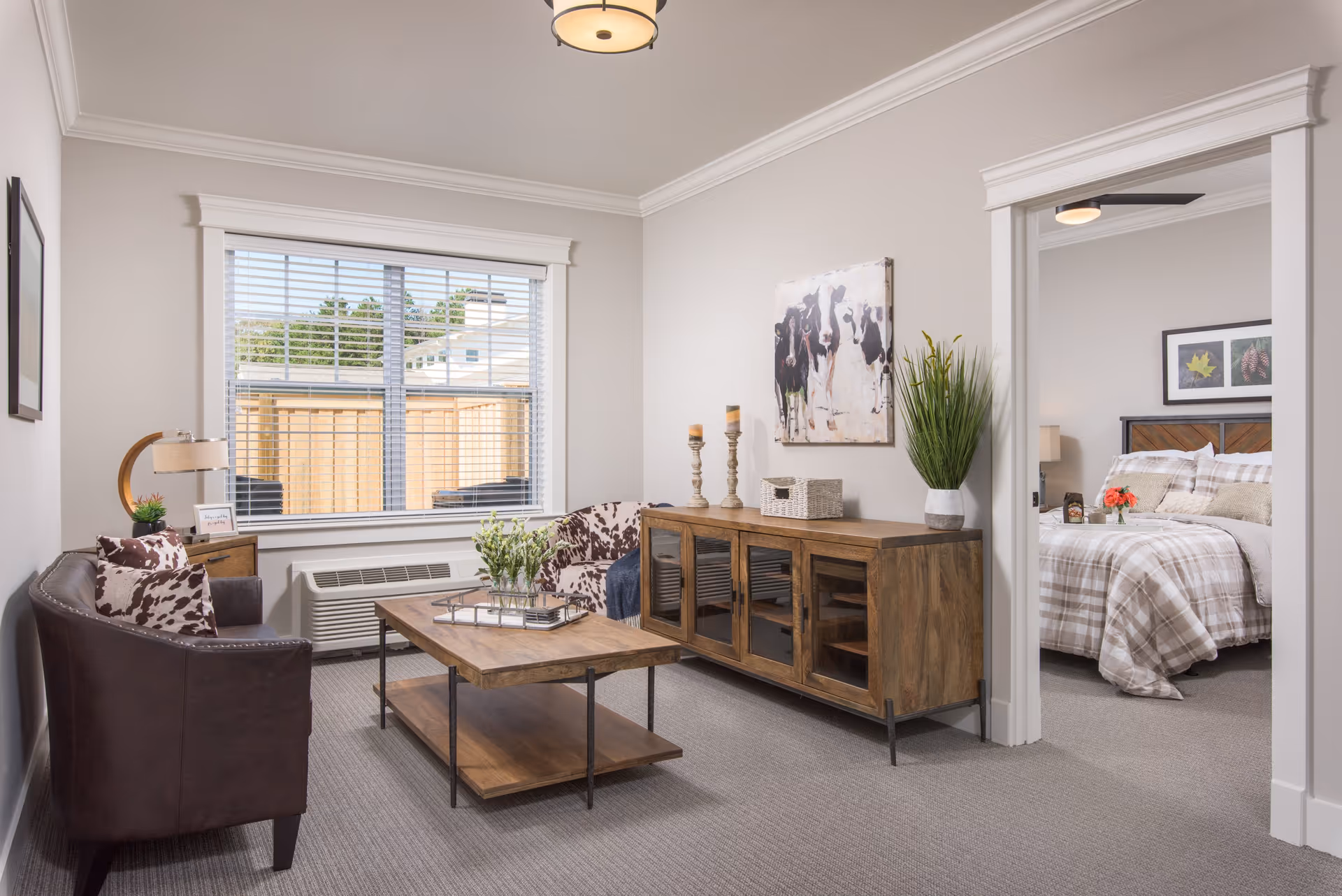 A cozy living room with a brown leather couch and a patterned armchair, a wooden coffee table with a plant centerpiece, a wooden sideboard with decorative items, and a large window with blinds. Through an open doorway, a bedroom with a bed covered in plaid bedding and framed artwork on the walls is visible.
