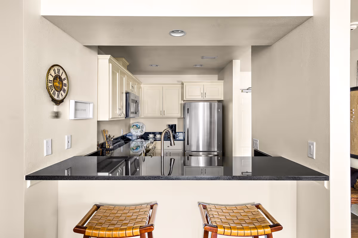 View of a modern kitchen with white cabinets, a stainless steel refrigerator, a microwave, and a black granite countertop with a sink and faucet. Two woven bar stools are positioned at the counter. A decorative wall clock and a small framed sign are mounted on the left wall.