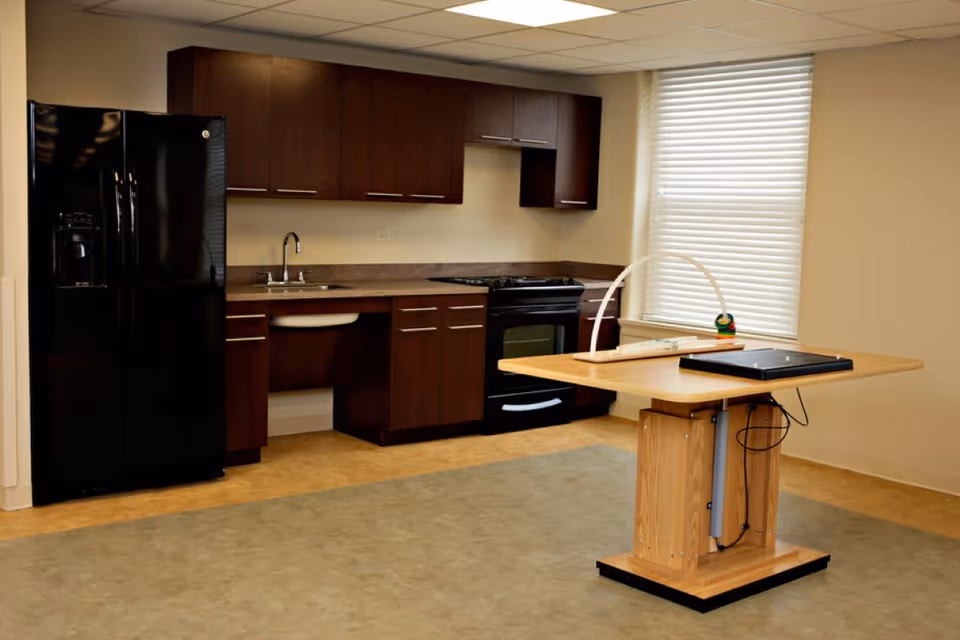 A kitchen area with dark wood cabinets, a black refrigerator, a black stove, and a sink. In front of the kitchen is a wooden adjustable table with a laptop and a small toy on it. A window with white blinds is on the right side of the room.