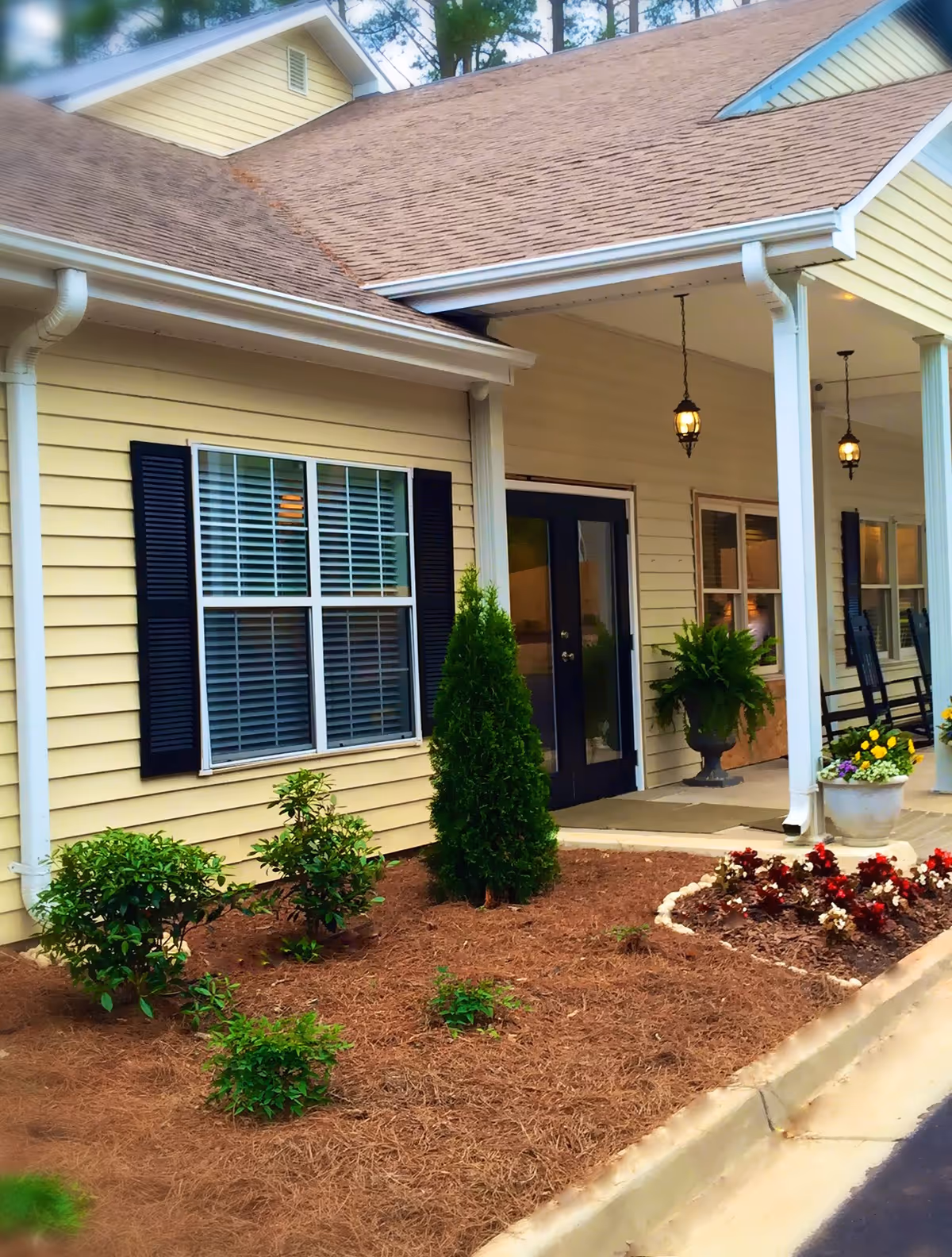 Front exterior view of a senior living community building with beige siding, black shutters, a covered porch with hanging lanterns, potted plants, and a landscaped garden bed with small shrubs and flowers.