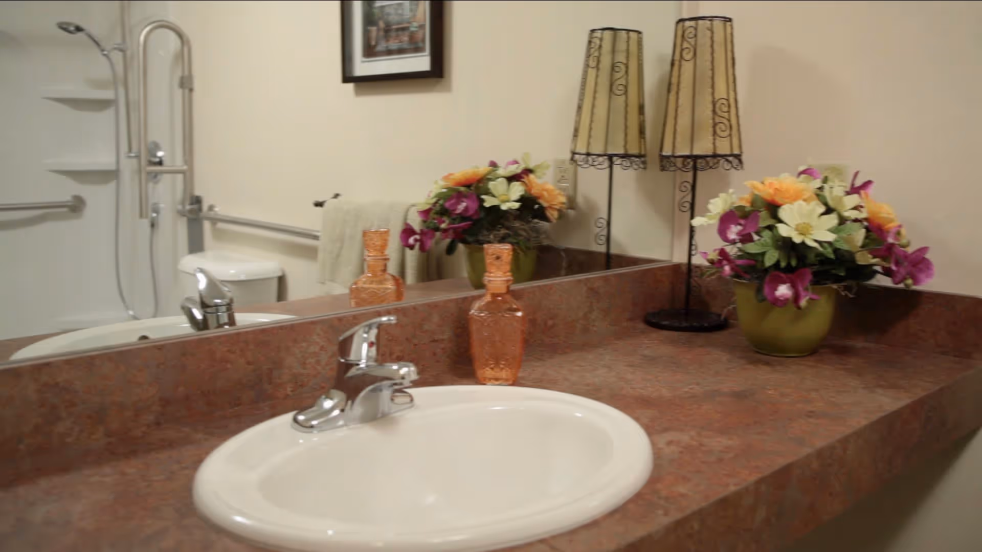 Bathroom countertop with a white sink and silver faucet. On the counter are a decorative glass bottle, a green vase with colorful flowers, and a lamp with a beige shade. In the background, a toilet, a towel hanging on a grab bar, and a shower with grab bars are visible. A framed picture hangs on the wall above the towel.