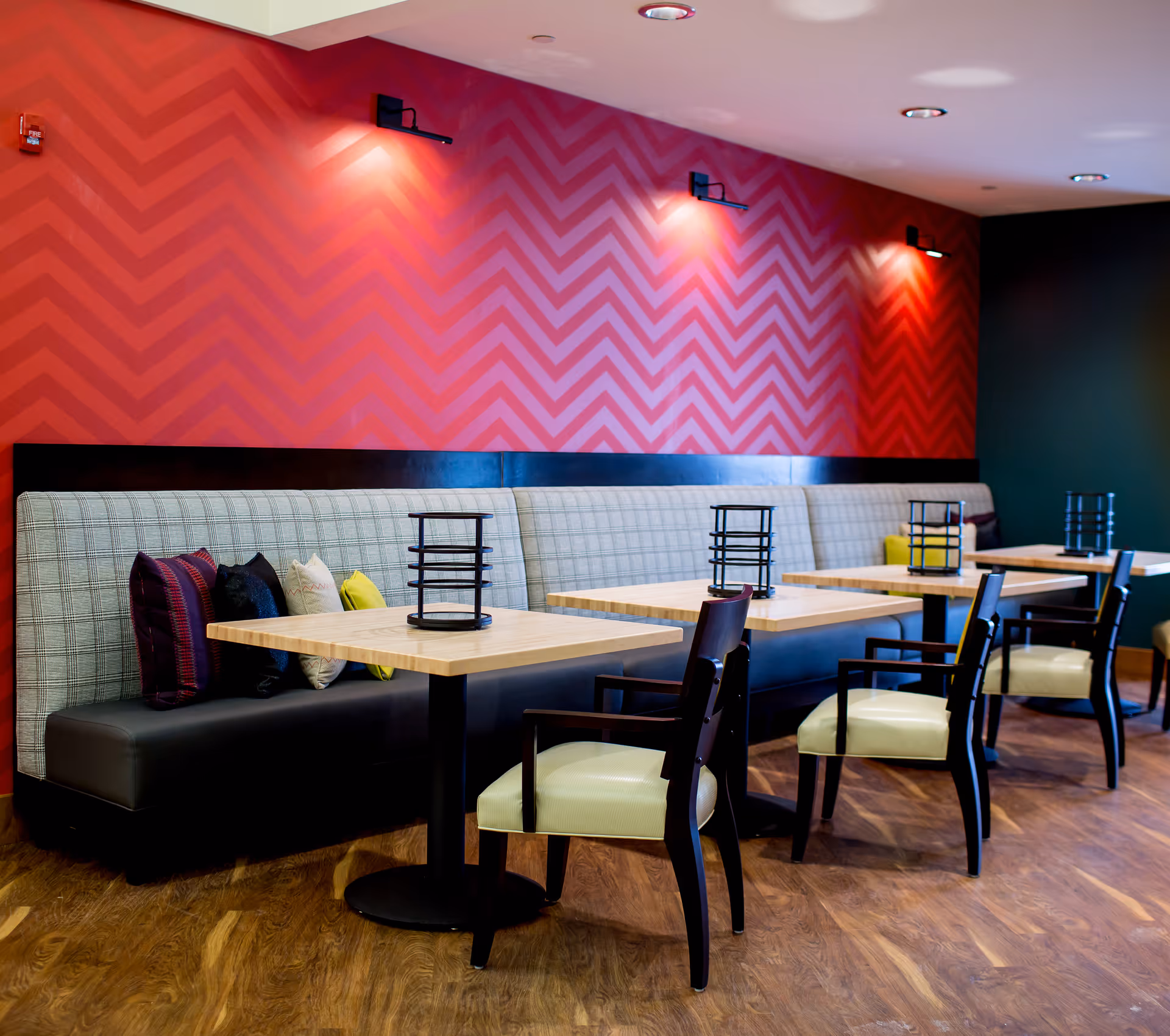 Interior view of a dining area with wooden tables, cushioned chairs, and a long bench seat with patterned pillows against a red chevron-patterned wall. The floor is wooden, and there are small black lantern-style centerpieces on each table.