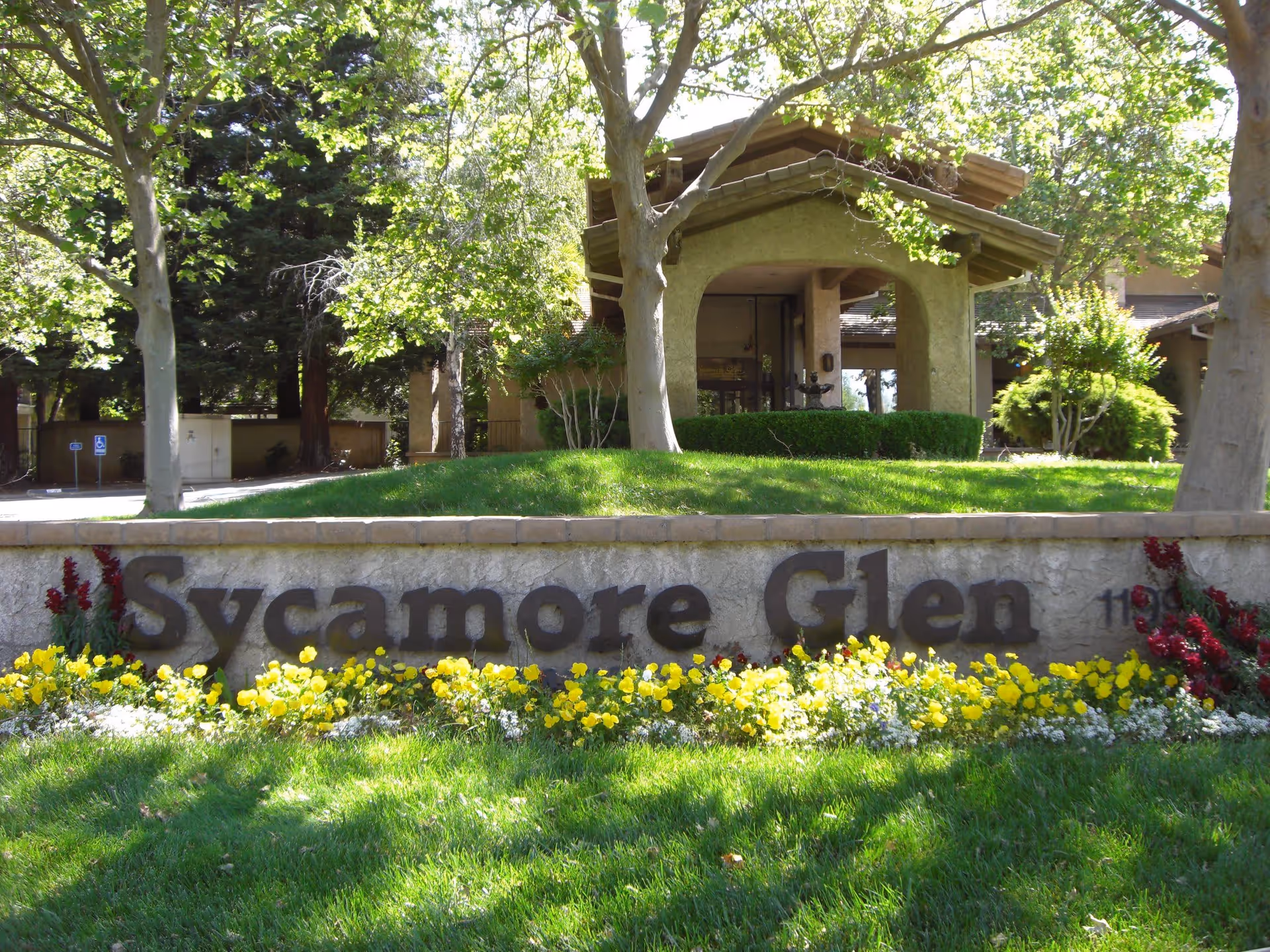 Entrance sign for Sycamore Glen Active Senior Community with a stone wall and colorful flowers in front, surrounded by green grass and trees, with part of the community building visible in the background.