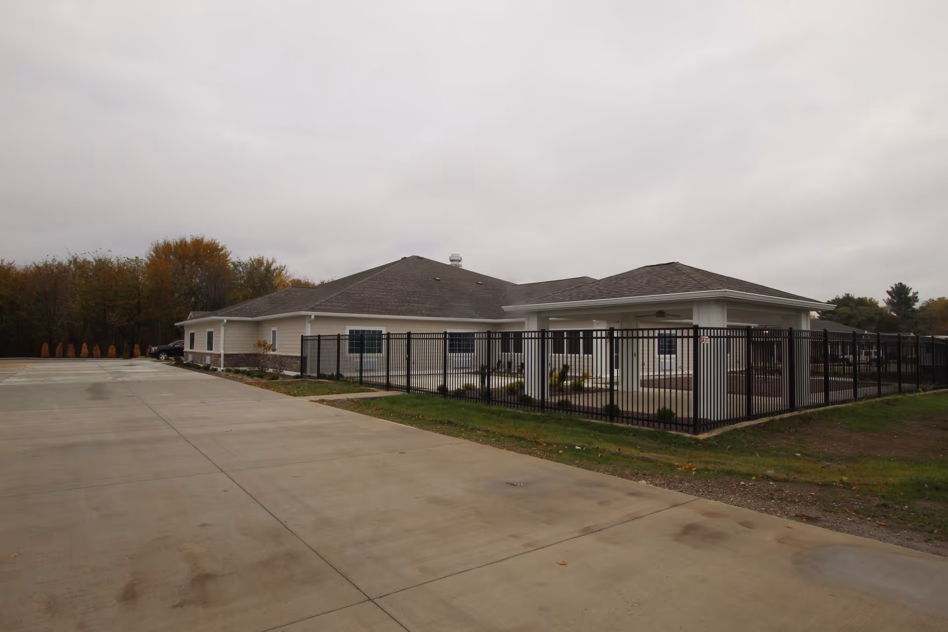 Single-story memory care building with a fenced patio and a wide concrete driveway under an overcast sky.