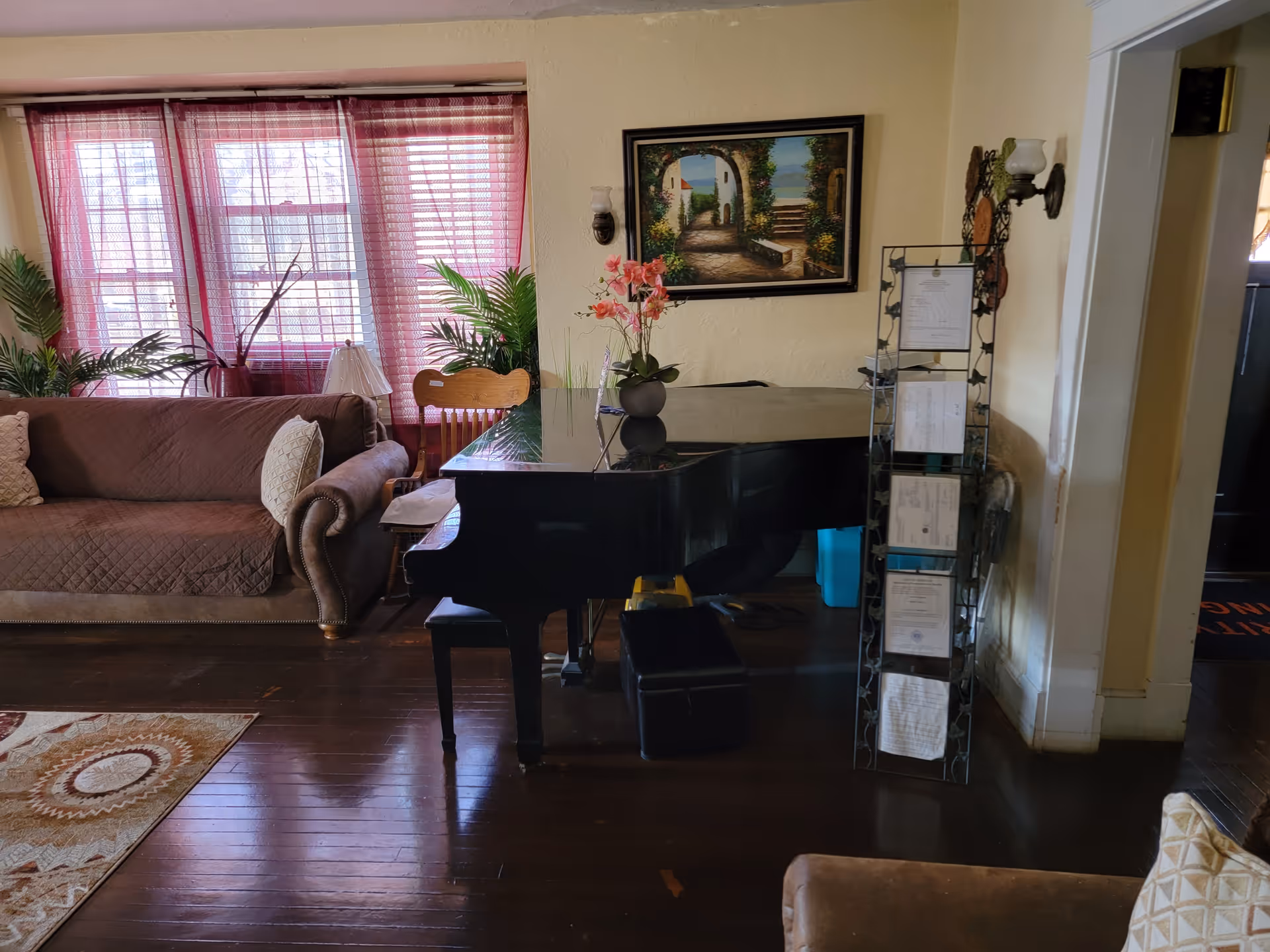 A cozy living room with a brown couch covered with a quilted cover and pillows, a black grand piano with a flower vase on top, a wooden chair, and a tall metal rack holding papers. The room has wooden floors, a large window with red curtains, green plants, and a framed painting on the wall depicting a scenic outdoor pathway.