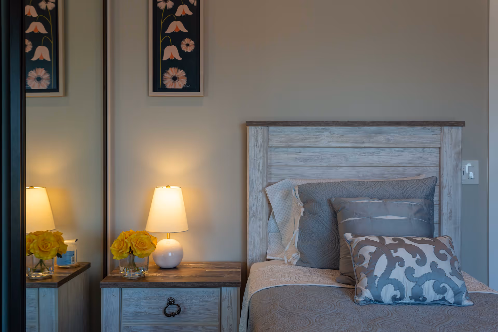 A cozy bedroom corner featuring a wooden headboard bed with decorative pillows in shades of gray and white. Next to the bed is a wooden nightstand with a white lamp and a small vase of yellow roses. A mirror on the left side reflects part of the nightstand and lamp. Above the nightstand, a framed floral artwork is hung on the wall.