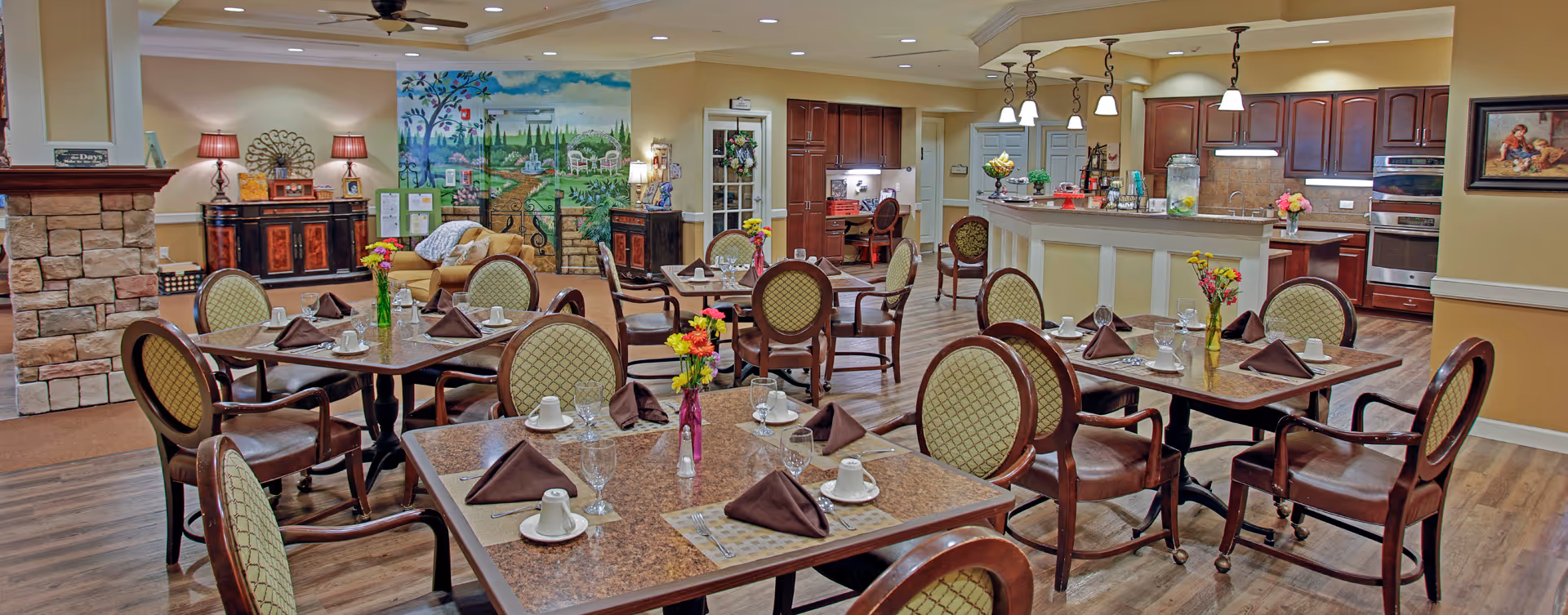 A dining room area in a senior living facility with several tables set with brown napkins, cups, glasses, and silverware. The room features wooden chairs with cushioned seats, a stone fireplace on the left, a colorful mural on the back wall, and a kitchen area with dark wood cabinets and pendant lighting on the right.