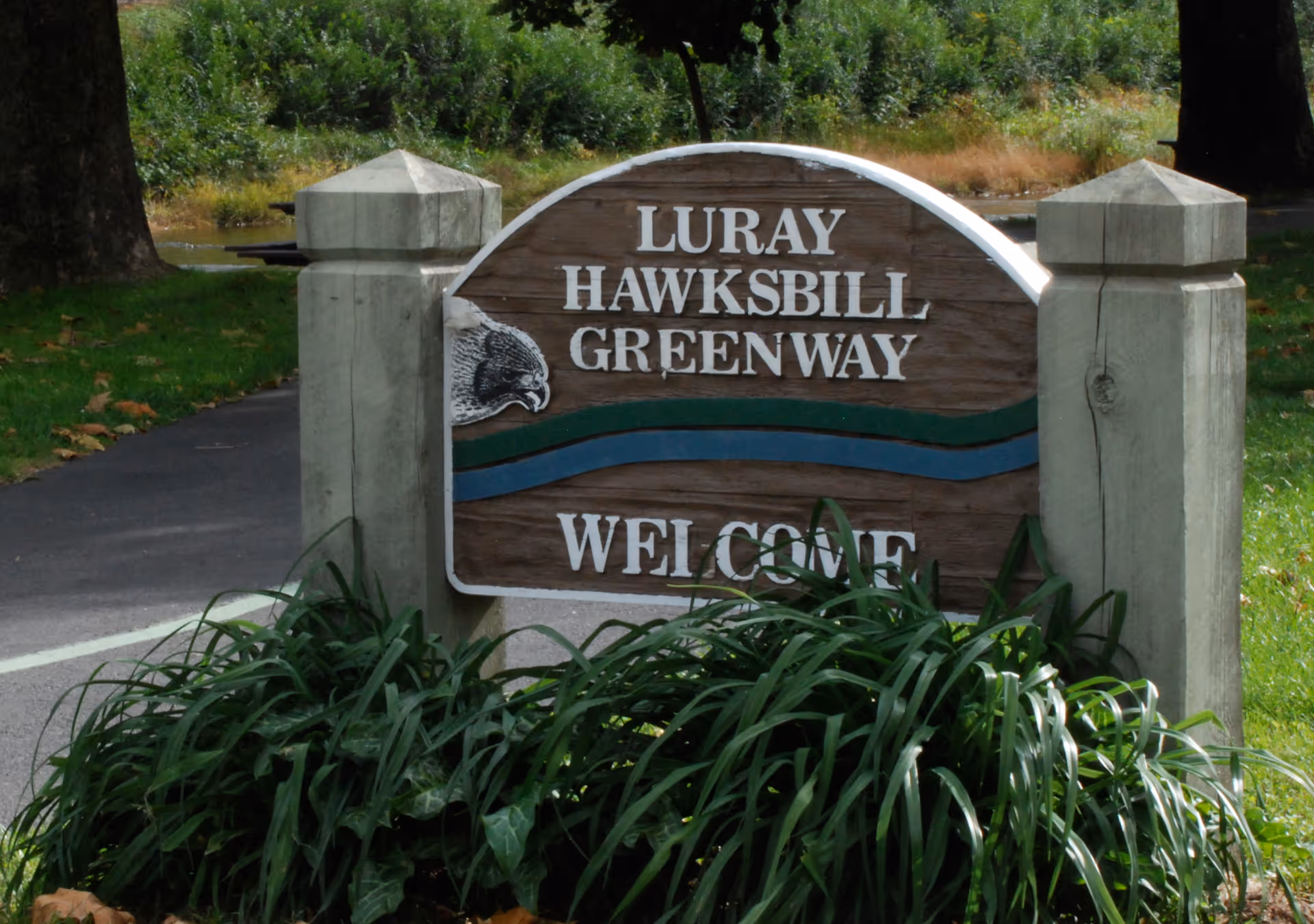 Wooden welcome sign reading Luray Hawksbill Greenway beside a paved path.