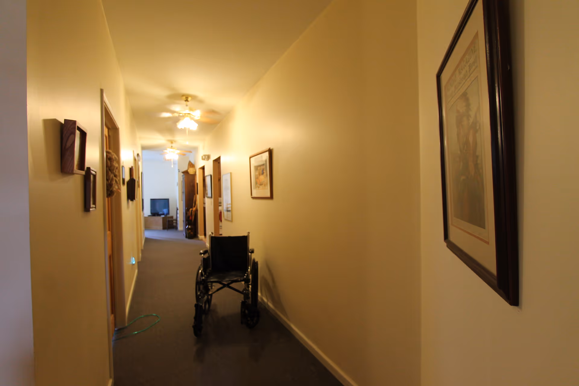 Long interior hallway in a senior living facility with a wheelchair parked in the center, framed pictures on the walls, and a TV visible at the far end.