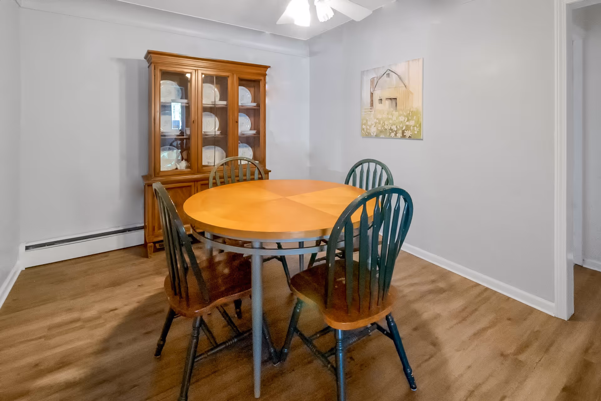 A dining room with a round wooden table surrounded by four wooden chairs with dark green backs and seats. Against the wall is a wooden china cabinet displaying white plates. A painting of a barn and flowers hangs on the light gray wall. The floor is wood, and a ceiling fan is visible above.