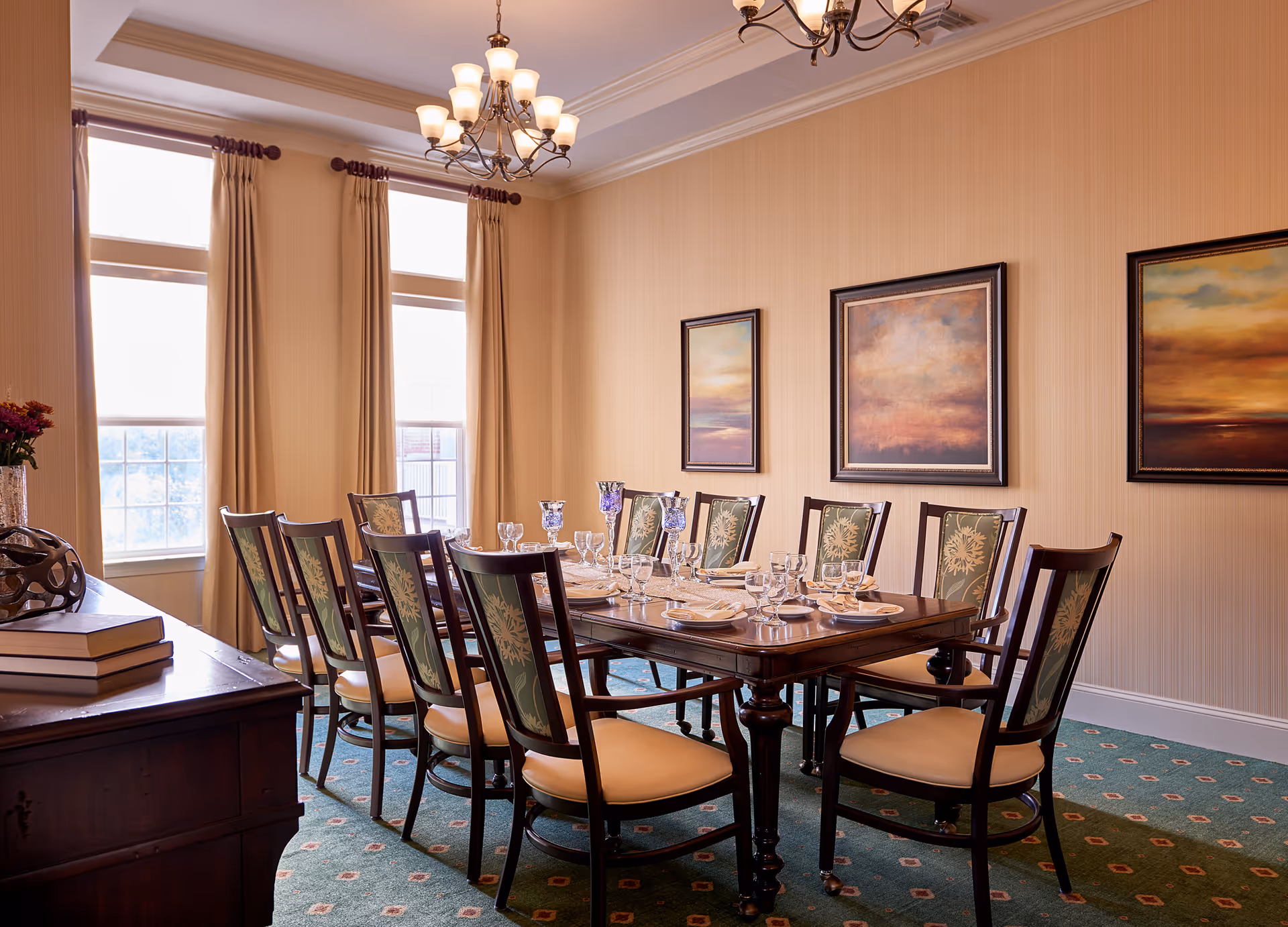 A formal dining room with a long wooden table set for a meal with plates, glasses, and napkins. The room has ten chairs with floral patterned backs and beige cushions. There are three large windows with beige curtains, three framed paintings on the wall, and two chandeliers hanging from the ceiling. A wooden sideboard with books and a decorative item is visible on the left side.