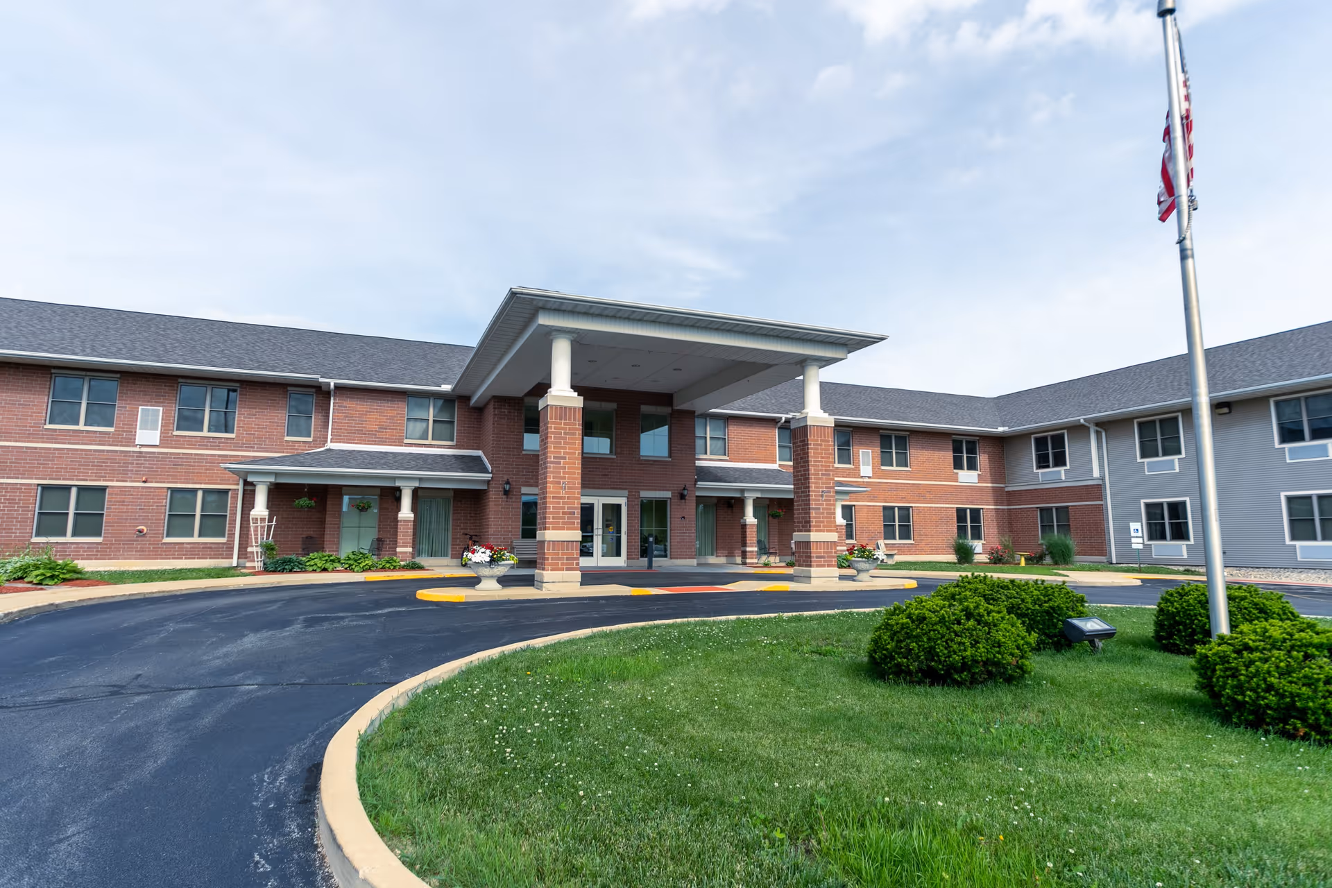 Front exterior view of a two-story senior living facility building with a covered entrance supported by columns, a circular driveway, green lawn, bushes, and an American flag on a flagpole.