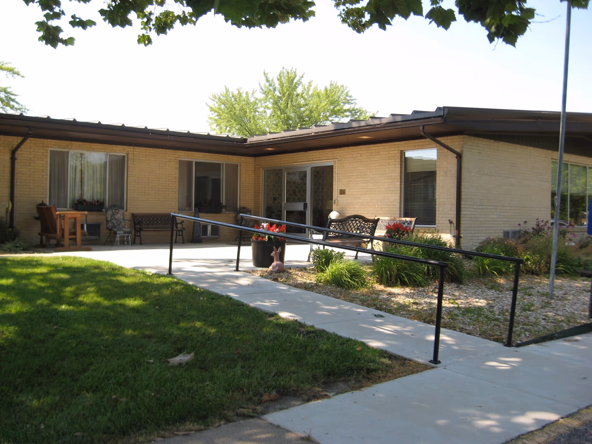 Exterior view of a single-story brick building with large windows and a glass door entrance. There is a concrete walkway with a black metal handrail leading up to the entrance, surrounded by green grass, plants, and flowers. Several benches and chairs are placed near the building's entrance under a shaded area.